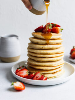 Pouring maple syrup over a stack of fluffy vegan pancakes and fresh strawberries