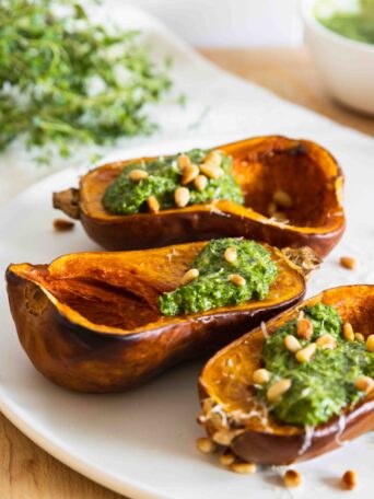 Roasted honeynut squash served on a white plate, topped with fresh pesto, toasted pine nuts, and vegan parmesan.