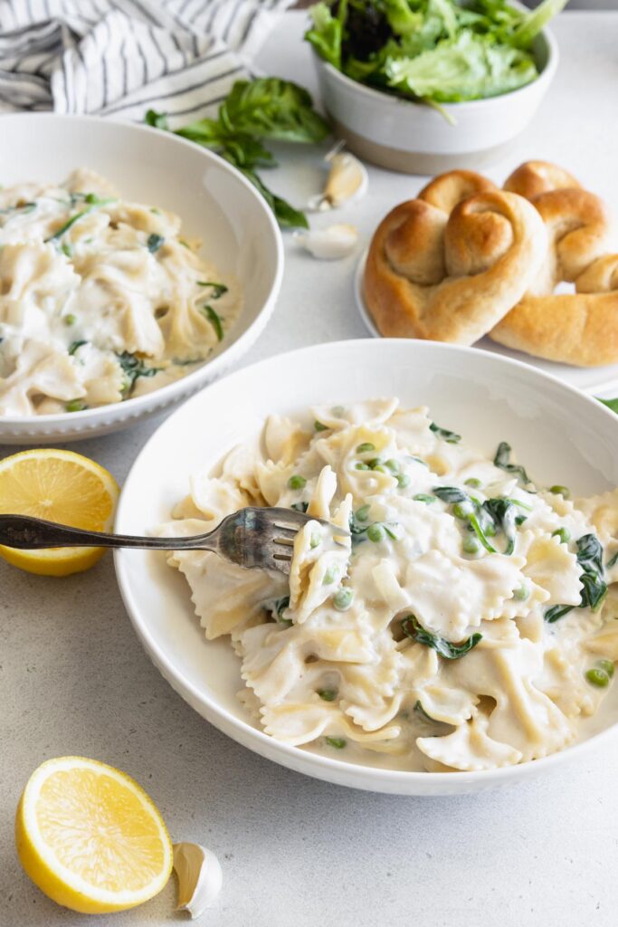 Creamy vegan pasta served with spinach and peas in a white bowl with fresh rolls